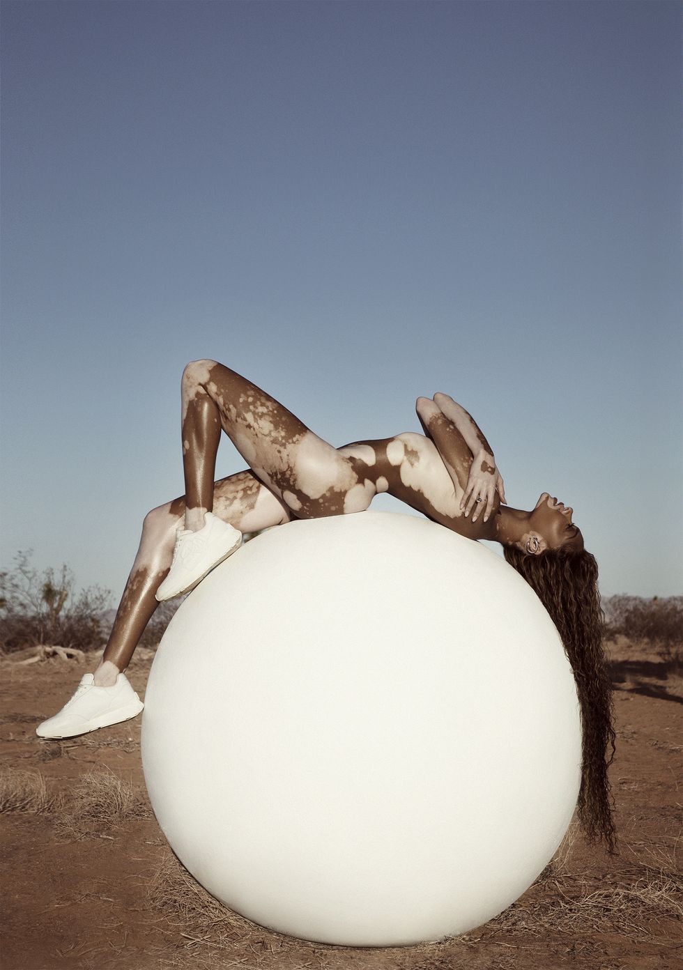 model winnie harlow lying bare on her back on big concrete white ball, head looking up at sky, arms crossed covering her upper body, on an open desert like landscape with blue sky